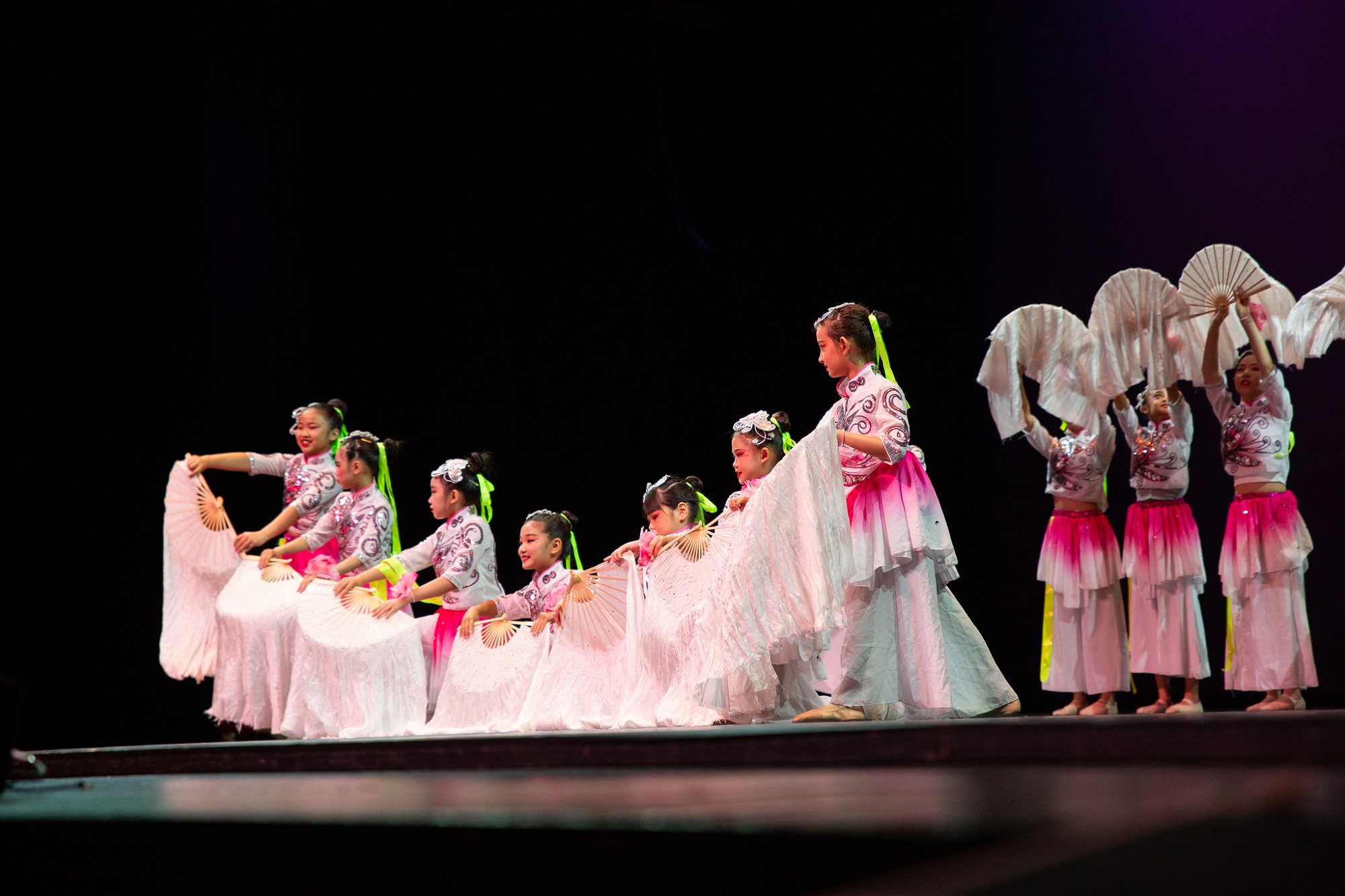 A group of young performers in traditional Chinese attire, holding white fans and dressed in pink and white costumes, are dancing on a stage under colored lighting.