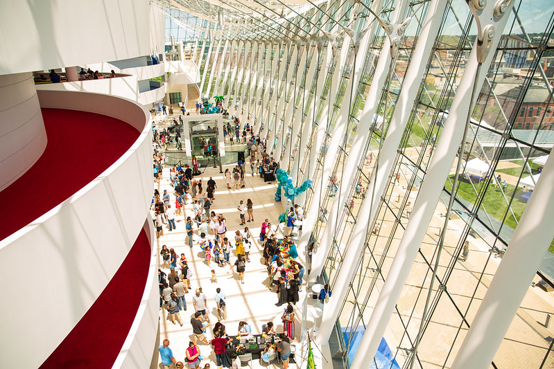 A crowd of people gather in a spacious, sunlit atrium with a high glass wall and red carpeted balconies, where signs highlight various Ways to Give.