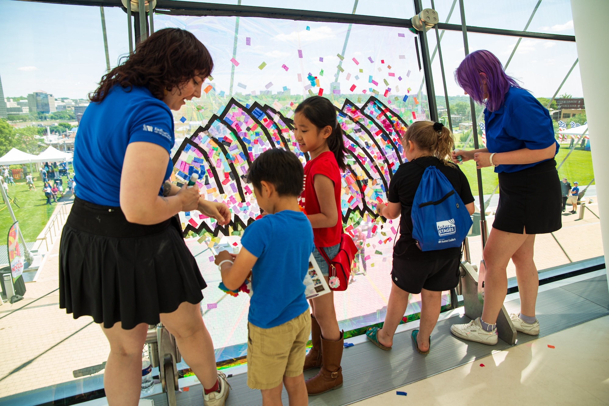 Children and adults interact with a colorful art installation near a window. The backdrop shows an outdoor event with green grass and tents, showcasing the vibrant community supported by nonprofit jobs in Kansas City.