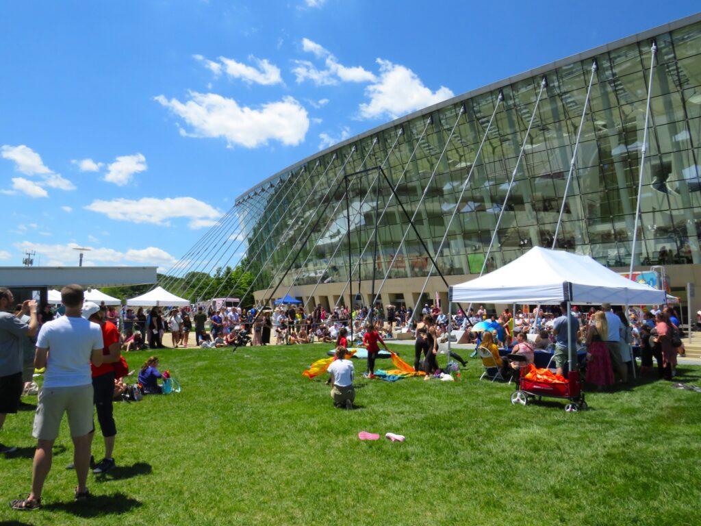 A crowd gathers outside a modern glass building on a sunny day. Several white tents, part of a Kauffman Center Festival, are set up on the green lawn, with people sitting or standing around.