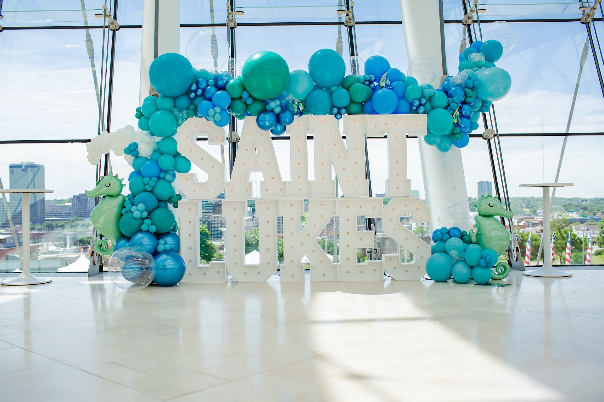 Large "Saint Lukes" sign decorated with blue and green balloons, two seahorse figures, and set against the backdrop of a glass-windowed room with a cityscape view, highlighting the influence of corporate sponsorships.