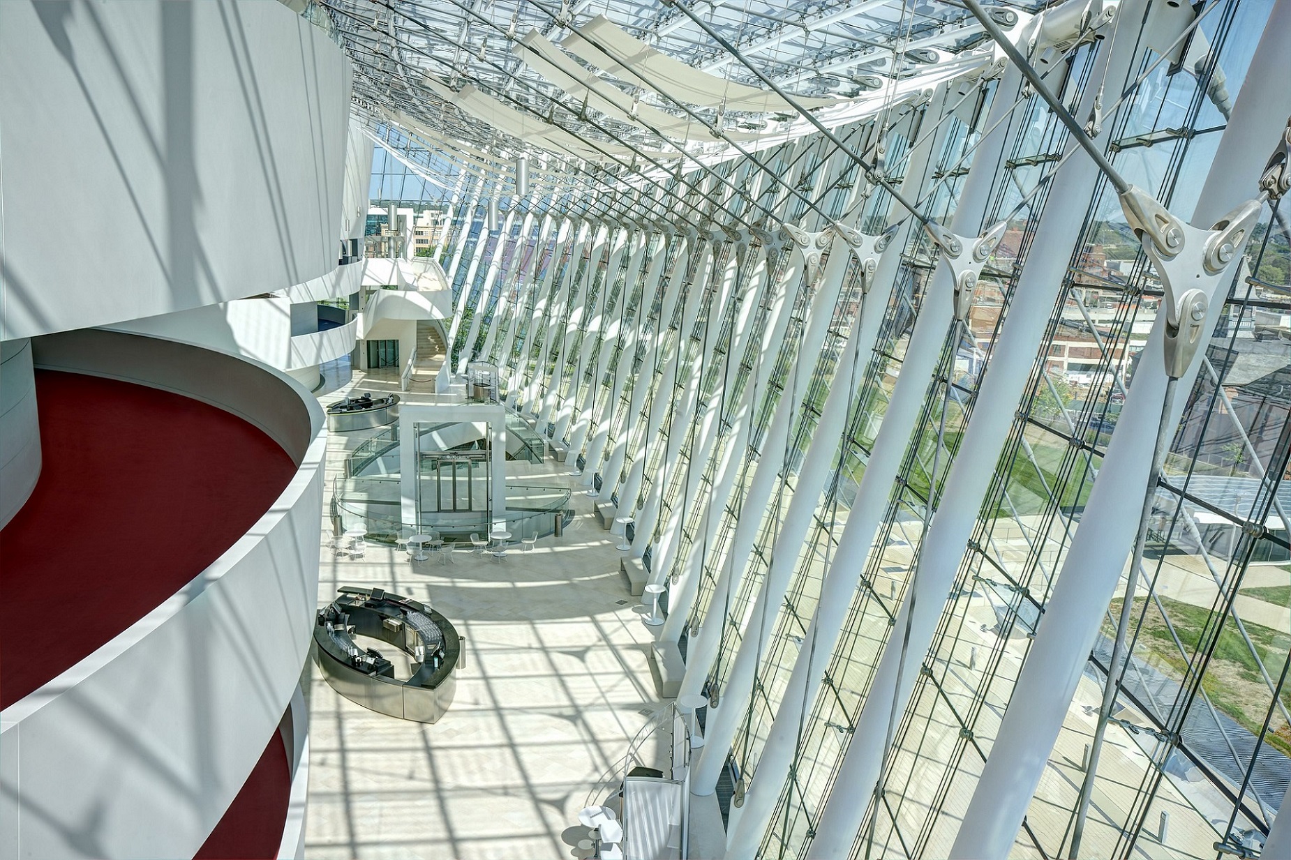 Brandmeyer Great Hall, a spacious modern building features a tall, curved glass facade with vertical support beams. Inside, a red-carpeted balcony overlooks a sunlit atrium with white tables. Photo by Steve Mohlenkamp Photography.