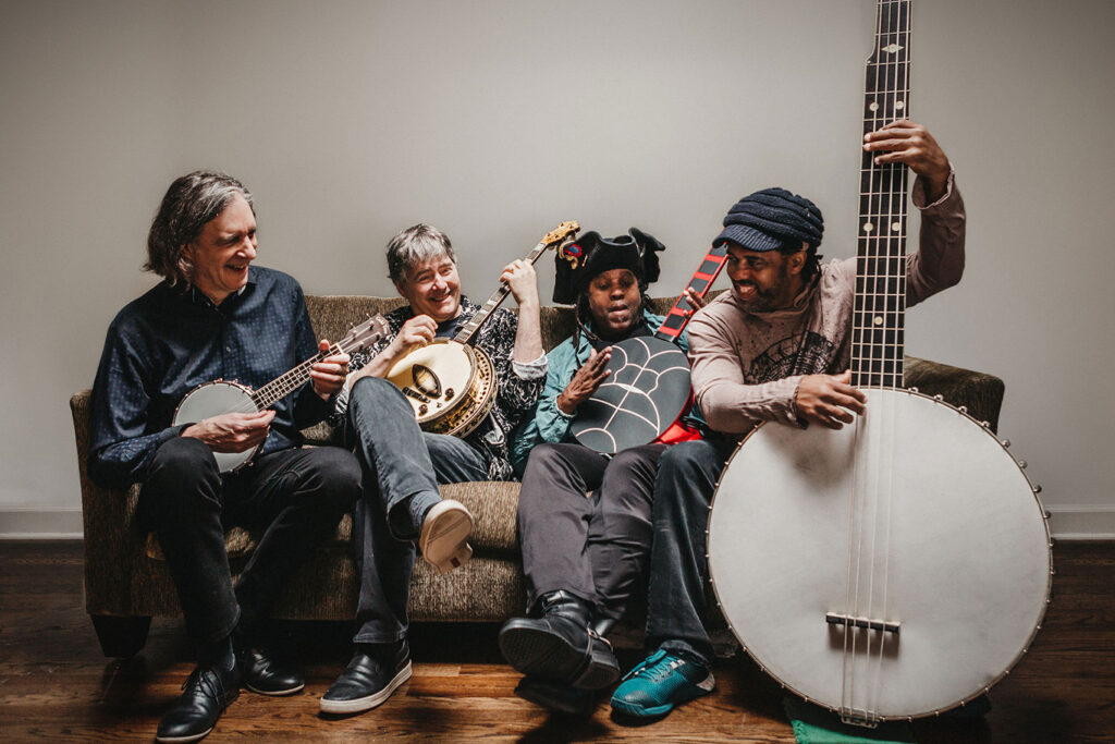 Four people sit on a couch playing different string instruments, including banjos and a bass. They appear to be enjoying the moment together, reminiscent of a performance you might see at Kauffman Center Presents.