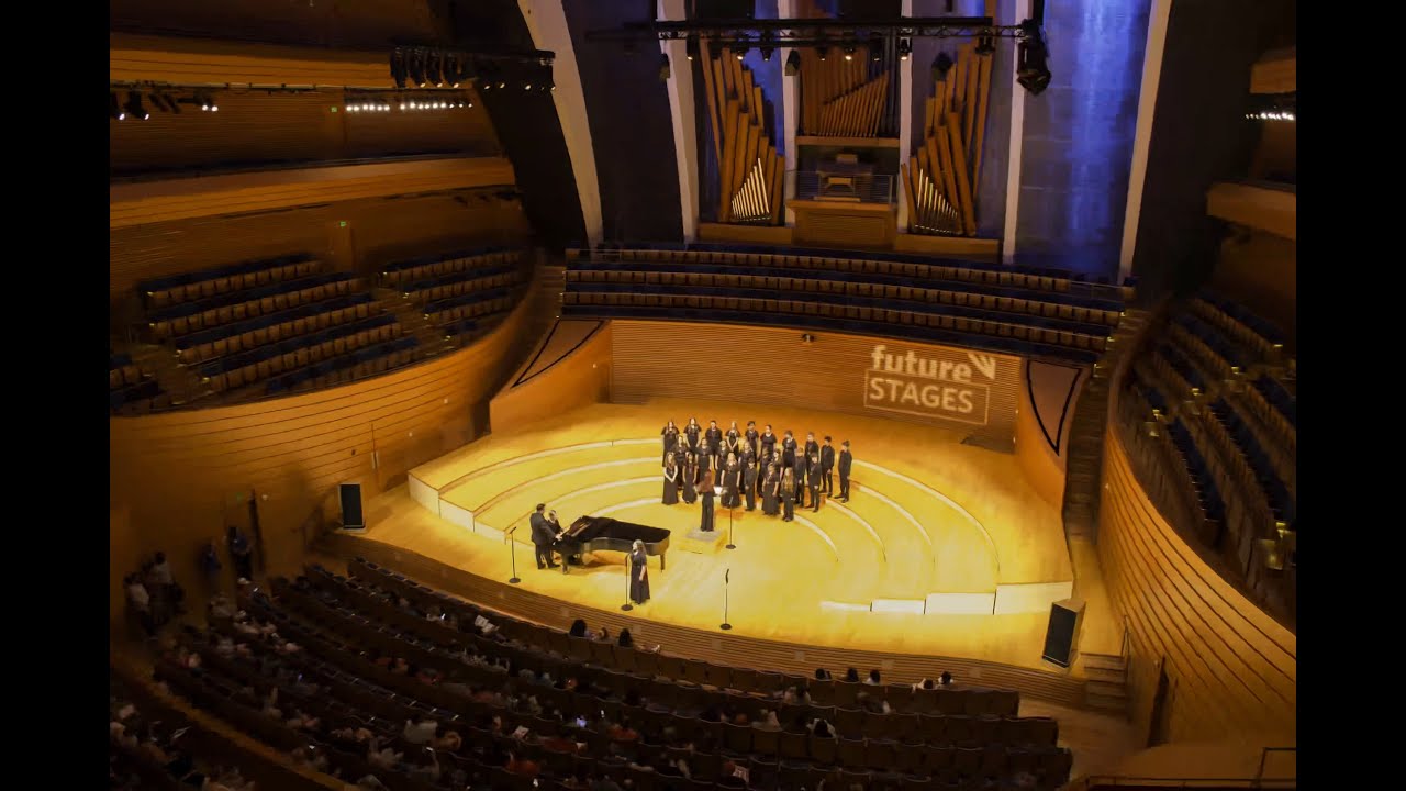 A choir performs on a well-lit stage in a modern concert hall with wooden interiors and an audience in attendance. The words "Future Stages Festival" are projected on the organ pipes in the background.