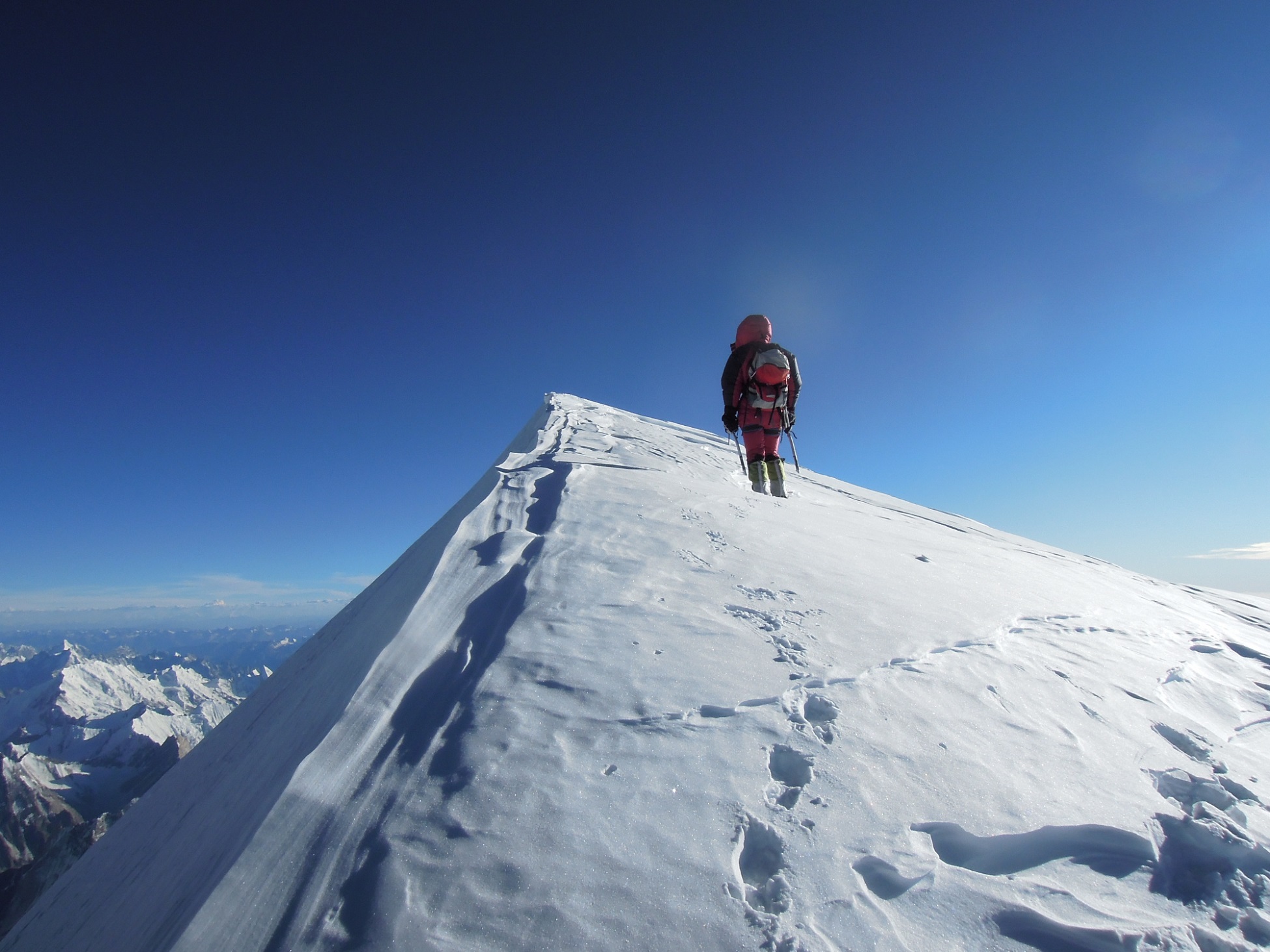 A mountaineer in red gear stands at the snowy peak of a high mountain under a clear blue sky. Footprints trail behind them along the snow-covered ridge. K2 Expedition –Photo by Gerlinde Kaltenbrunner