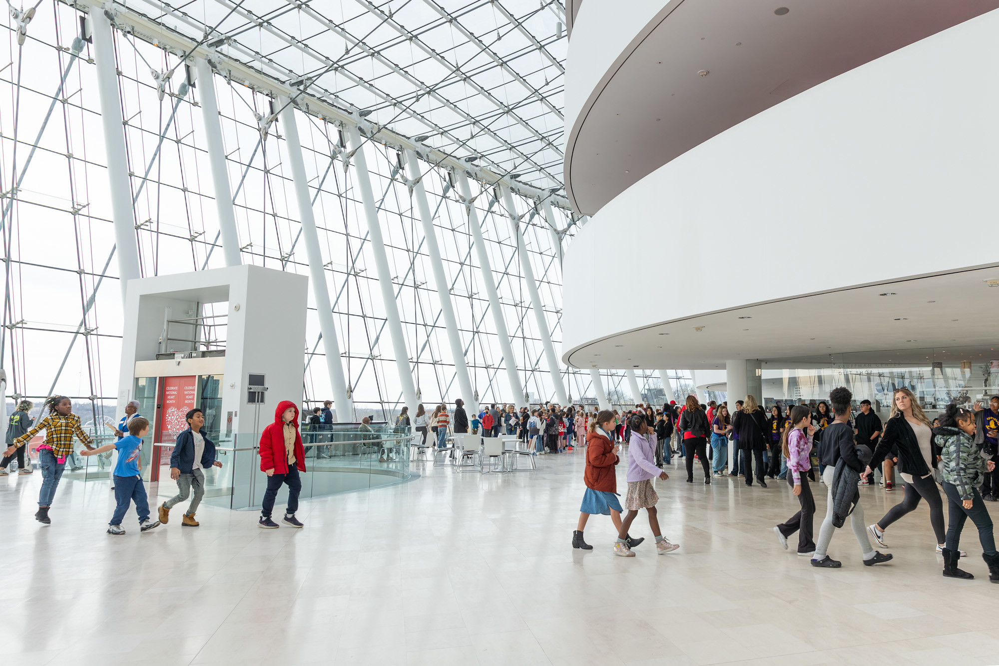 A large group of people walk through Brandmeyer Great Hall, a modern, spacious indoor lobby with glass walls and a curved architectural feature. | Photo by Lifted Logic.