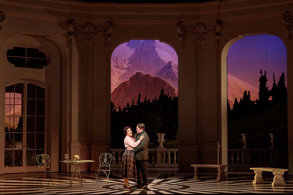 Two actors embrace on a stage set designed to resemble an elegant room with large arched windows overlooking a mountainous landscape. As part of the Performing Arts Sampler, a small table and chairs are positioned to the side, enhancing the intimate ambiance.