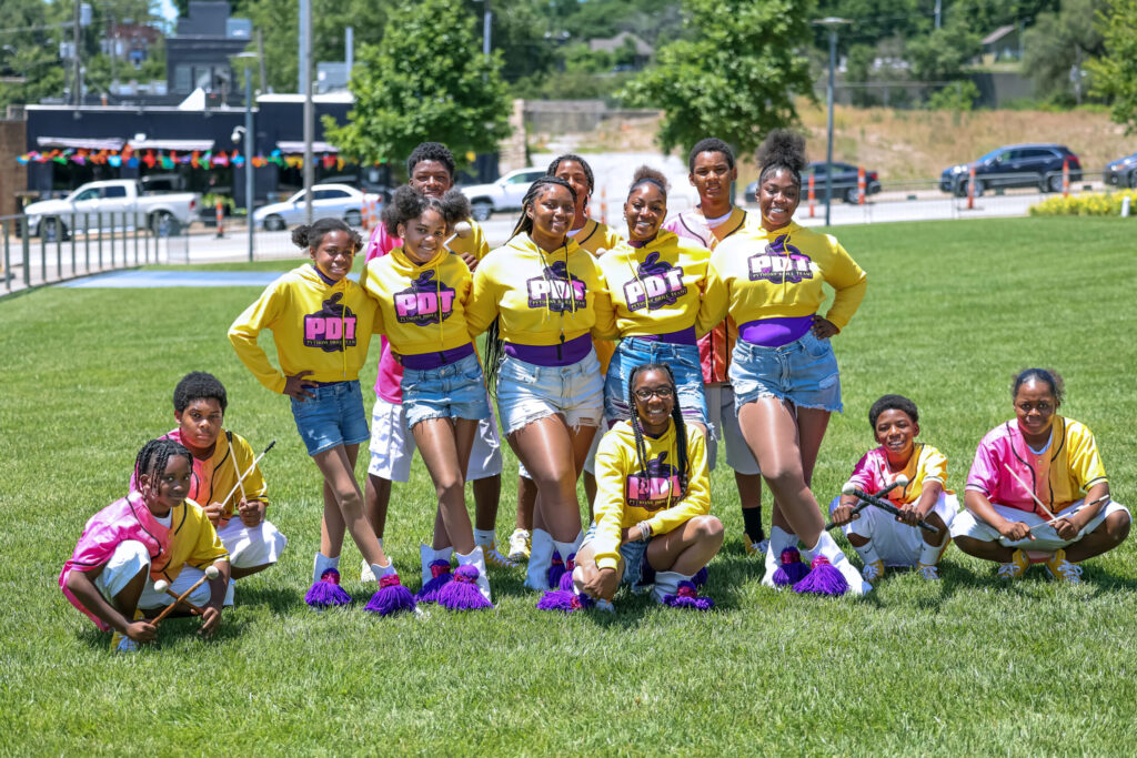 A group of kids wearing matching yellow and pink outfits pose on a grassy field, with some kneeling and others standing.