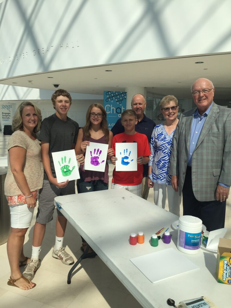 A group of people stands behind a table displaying handprint crafts. Three children hold their handprint artworks. The setting appears to be indoors with natural light.