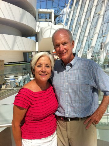 Two people standing close together and smiling inside a modern architectural building with large windows and curved structures.