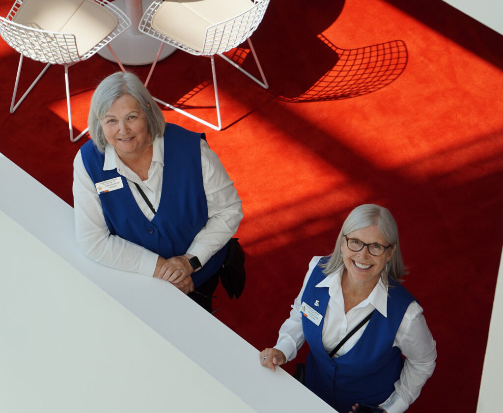 Two women in blue vests and white shirts are standing indoors on a red carpet near white chairs, smiling up at the camera, exemplifying the vibrant volunteer opportunities in Kansas City.