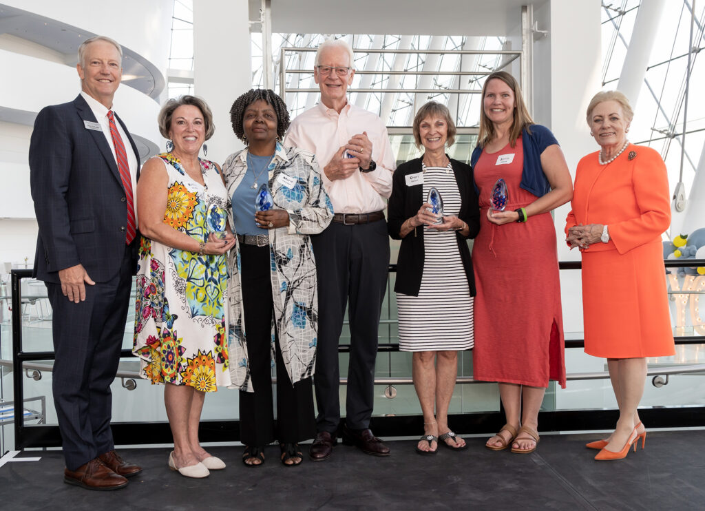 A group of seven people stand in a row indoors, each holding a small trophy. Five women and two men smile for the camera in formal attire, celebrating their achievements in volunteer opportunities in Kansas City.