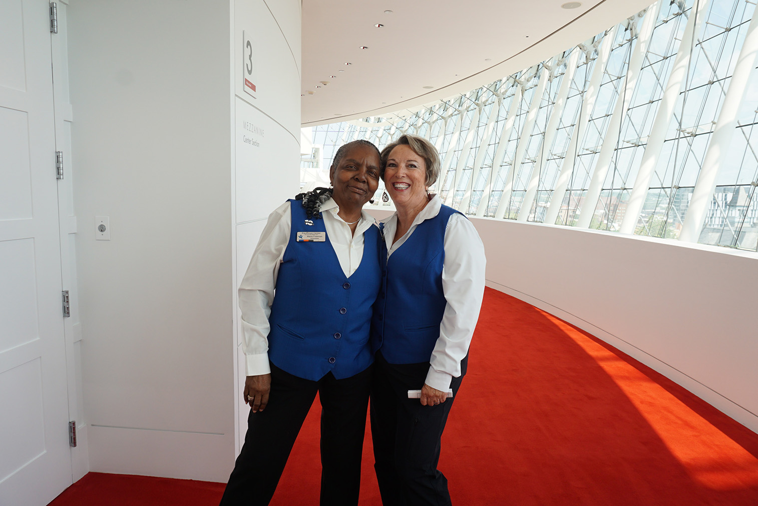 Two women wearing blue vests stand and smile in a brightly lit hallway with red carpet and large windows, embodying the spirit of volunteer opportunities in Kansas City.