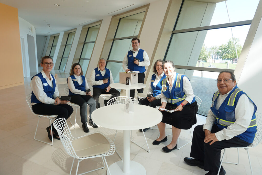 A group of seven people wearing blue uniforms with yellow vests sit and stand around small tables in a brightly lit room with large windows, discussing volunteer opportunities in Kansas City.