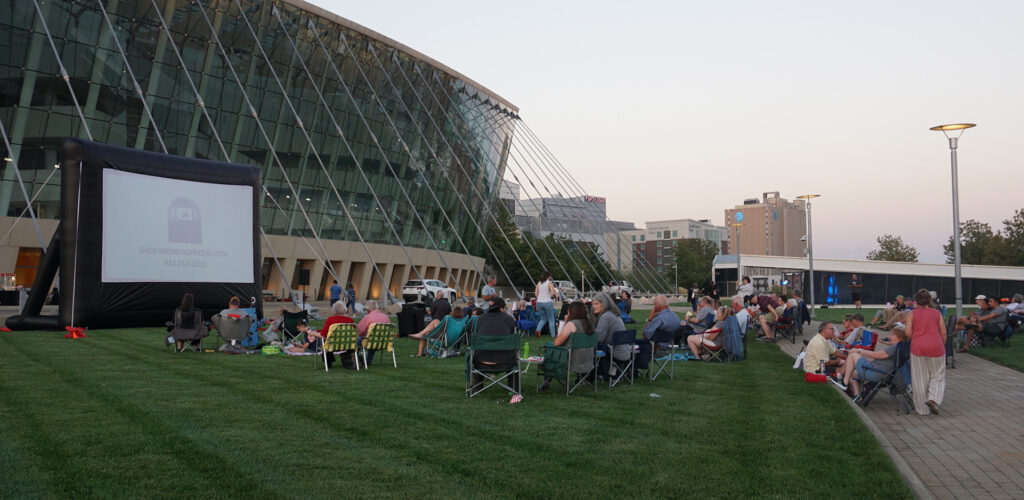 People are seated on a lawn watching an outdoor movie on a large inflatable screen near a modern glass building in the evening, enjoying one of the many volunteer opportunities in Kansas City.