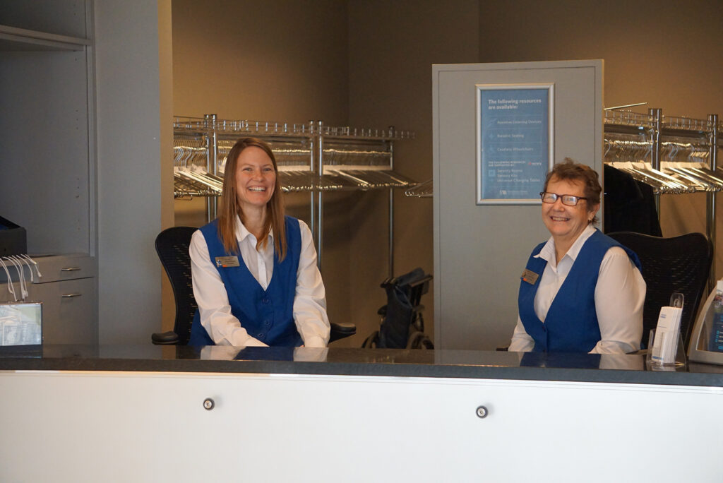 Two women in blue vests sit behind a Coat Check counter, smiling.