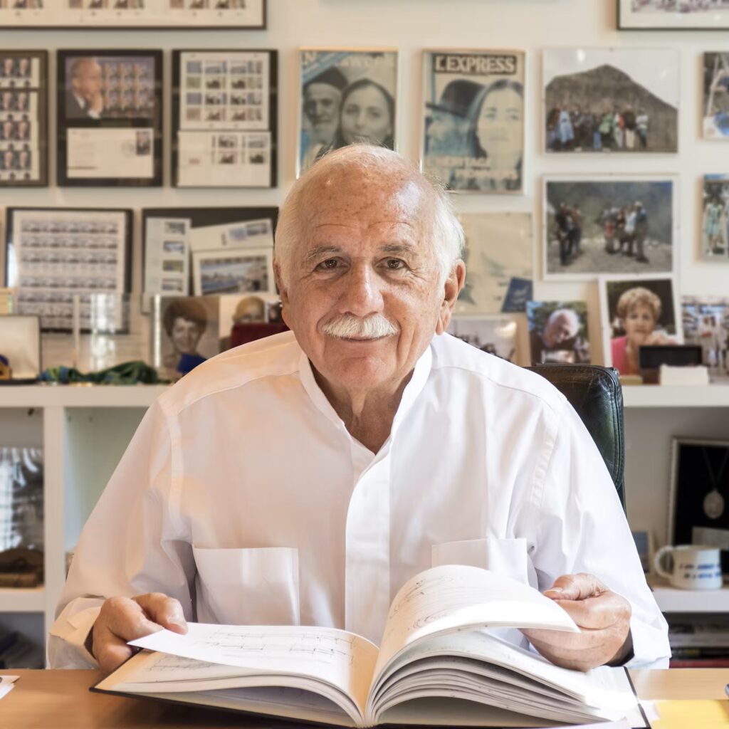 An elderly man with a white mustache, wearing a white button-down shirt, sits at a desk holding an open book. The wall behind him is covered with framed photos and media, including magazine covers and documents, creating a collage-like background.