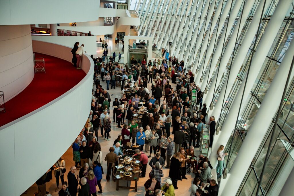 A large crowd of people mingle and socialize in an atrium with floor-to-ceiling windows. The space, belonging to a renowned Performing Arts Nonprofit in Kansas City, features modern architecture with curved glass walls and an upper balcony. Guests appear to be dressed for a formal or semi-formal event.