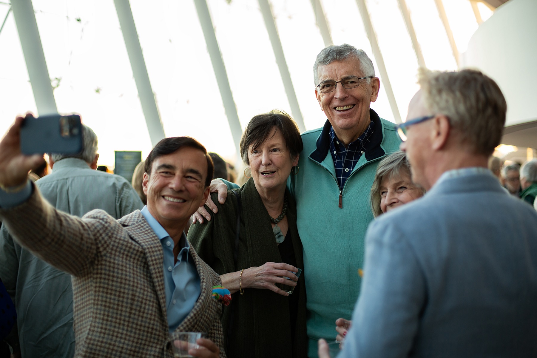 A group of five people, are gathered together at an indoor event hosted by a Performing Arts Nonprofit in Kansas City. One man in a plaid jacket is taking a selfie with a smartphone, while the others, smiling, pose for the photo. The background includes bright, tall windows, suggesting daytime.