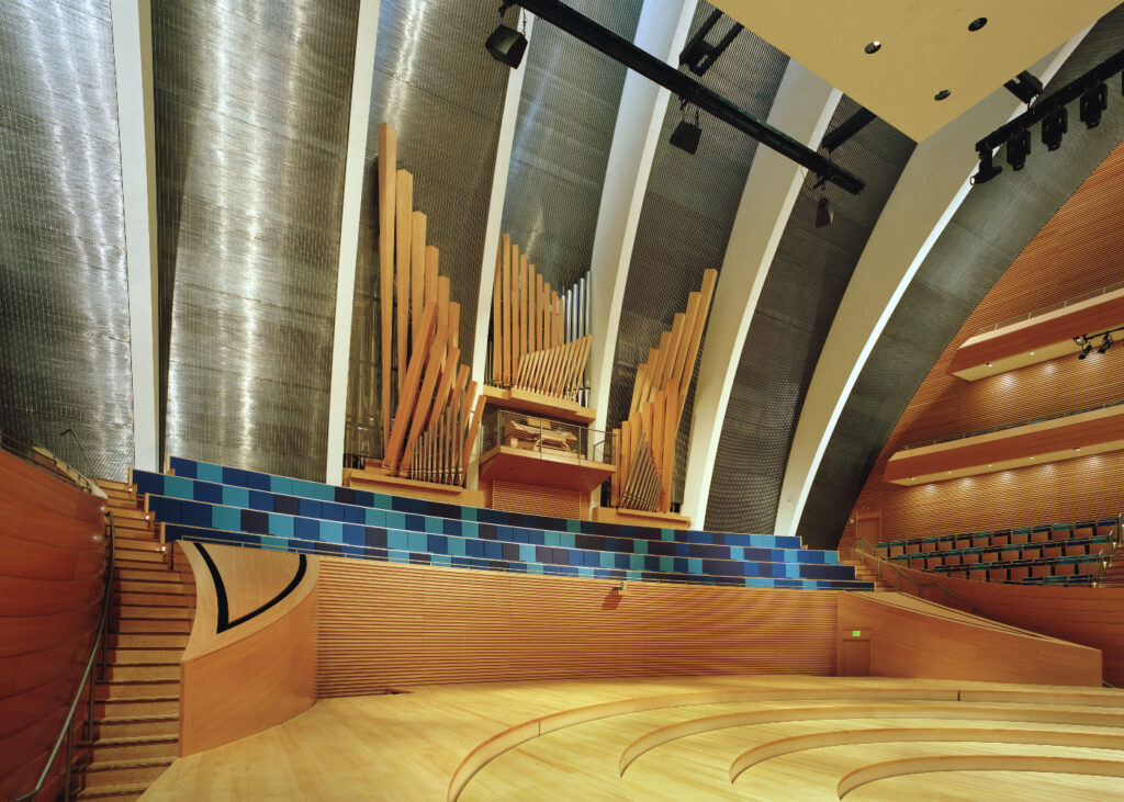 A modern concert hall featuring sleek architectural design with curved wooden seating areas and walls. The focal point is the Julia Irene Dennie-Kauffman Casavant Organ, boasting both wooden and metallic pipes, situated at the back of the stage under dramatic ribbed ceiling structures.