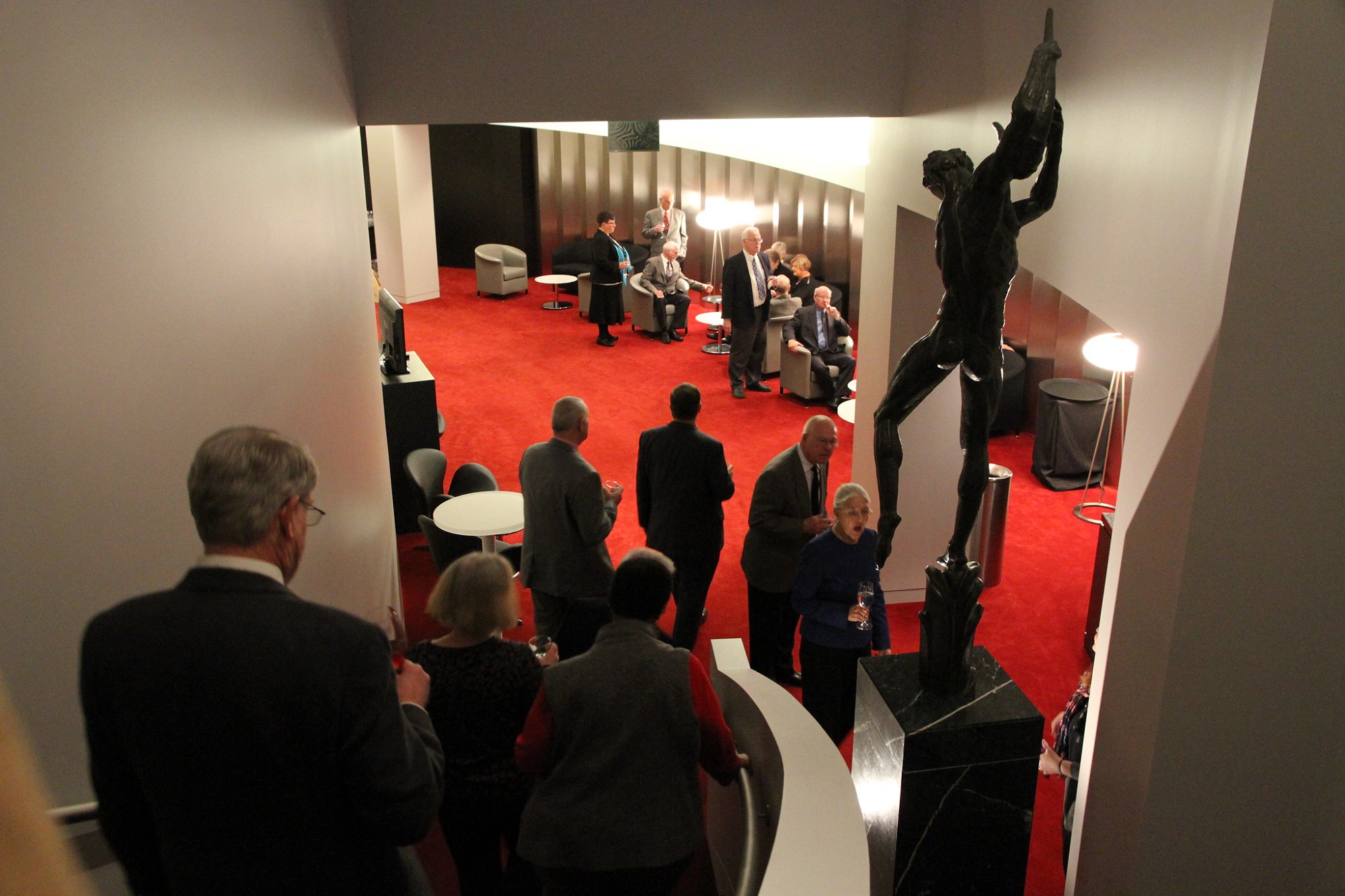 A group of people are mingling and sitting at tables in the Concert Hall Reception Suite with red carpeting and a tall sculpture. Some are standing on a staircase leading to the area.