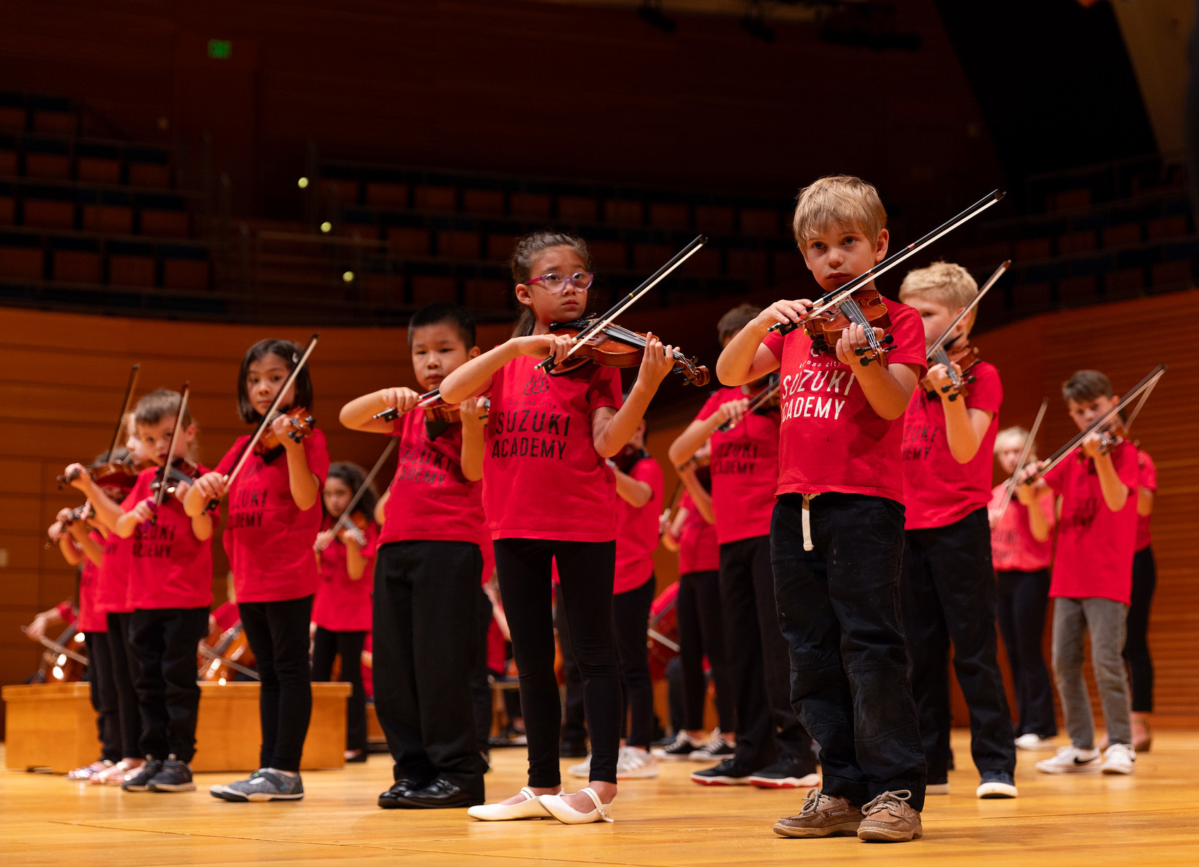 A group of children wearing red "Suzuki Academy" shirts perform on stage with violins. They stand in a row, each child focused intently on their instrument. The wooden stage and rows of seats in the background suggest a large auditorium or concert hall, part of the Future Stages Festival.