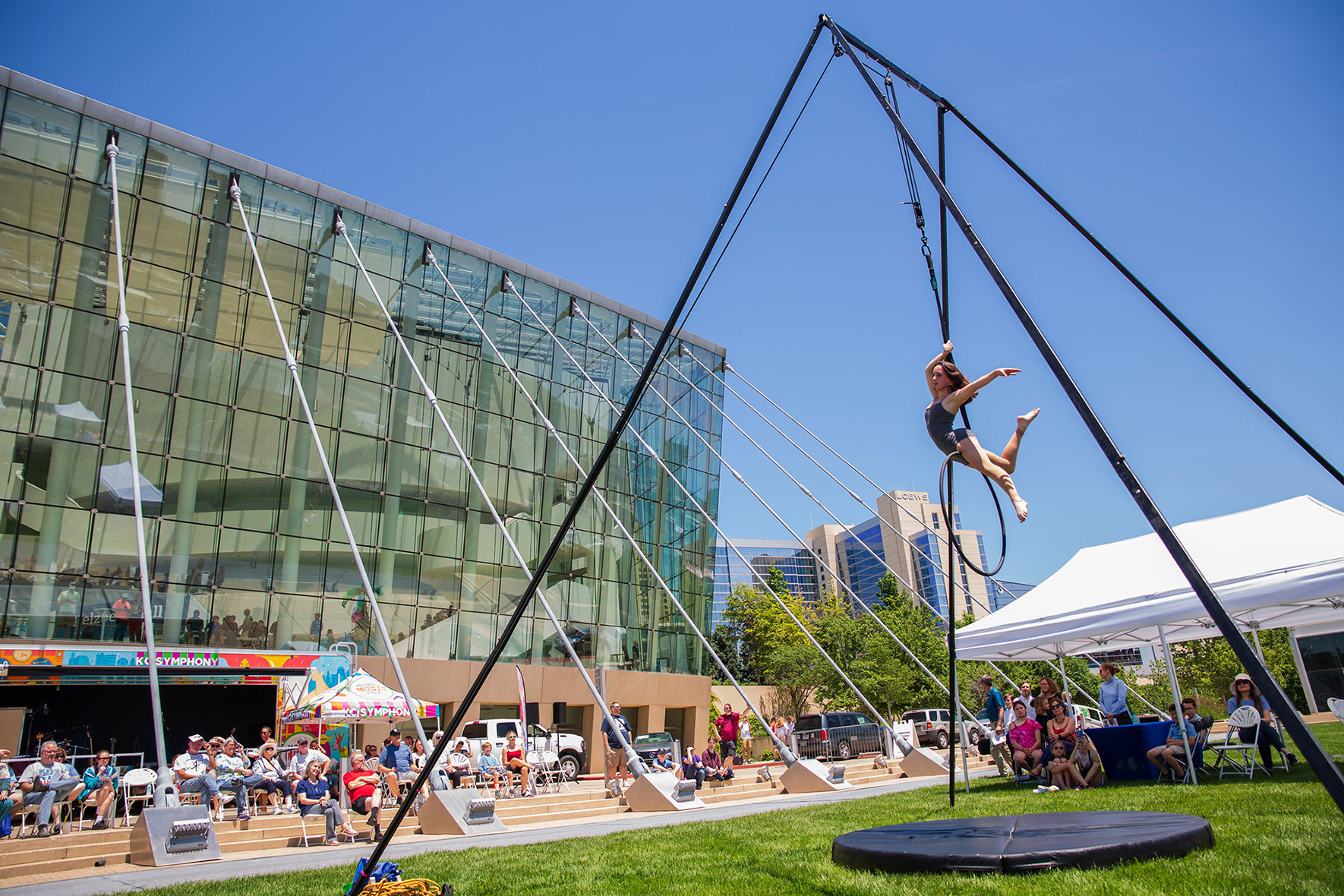 At the Future Stages Festival, a performer gracefully balances on a hoop suspended by a tall structure. Spectators sit under tents, watching the mesmerizing act in front of a modern glass building. The sky is clear and blue, adding to the magic of this sunny outdoor event.