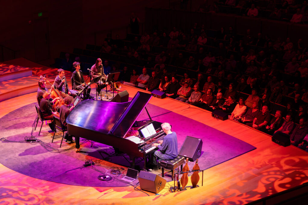 Musicians perform on stage with a grand piano, string instruments, and a guest singer while an audience watches in a dimly lit venue. In an effort to support the arts, patrons are introduced to various Ways to Give throughout the evening.