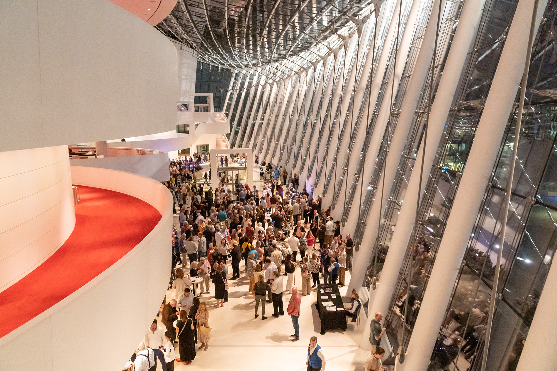 People gather in a spacious, modern atrium with large windows, white walls, and red carpeted balconies. Tables and displays by Kauffman Center for the Performing Arts in Kansas City are set up below.