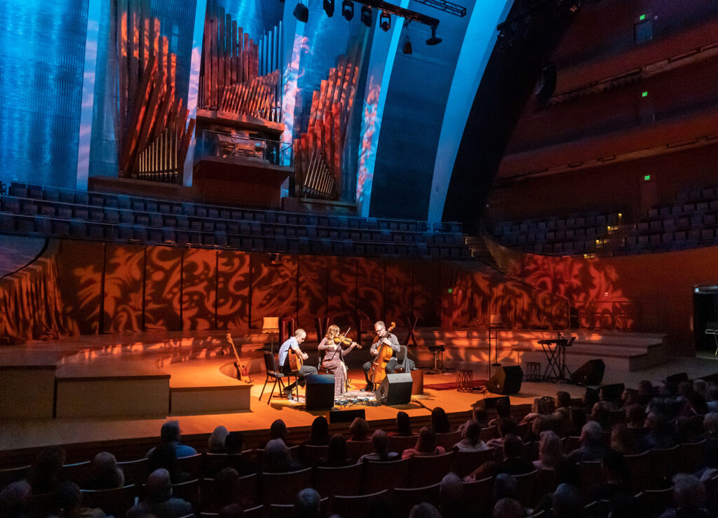 A string quartet from Kauffman Center for Performing Arts in Kansas City performs on stage in a dimly lit concert hall, with an illuminated organ in the background and an audience seated in front.