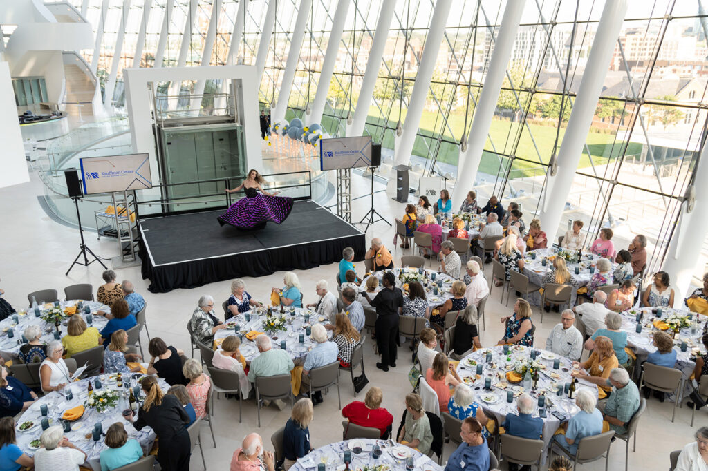 A performer in a purple dress dances on a stage in front of an audience at a volunteer banquet inside a modern, glass-walled building with views of Kansas City's skyline.