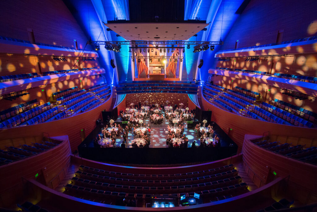 A wide-angle view of a large, modern concert hall at the Kauffman Center with a central stage set up for a formal event. Round tables and chairs are arranged on the stage, surrounded by dramatic lighting, available for rent.