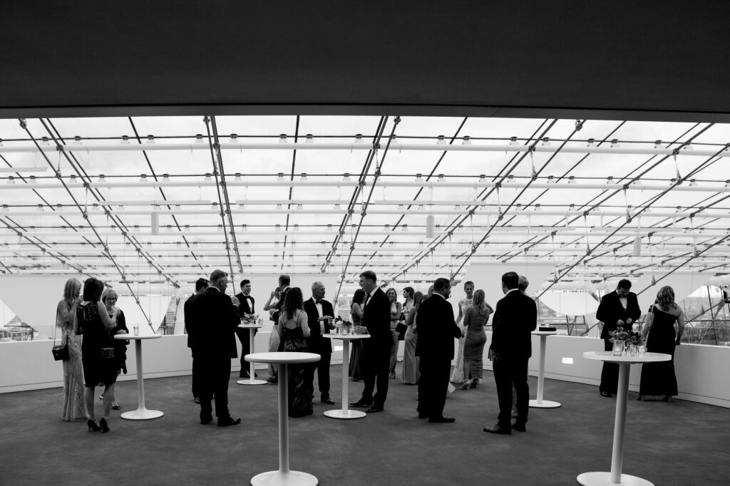 Black and white image of people in formal attire socializing around high tables in a glass-roofed event space.