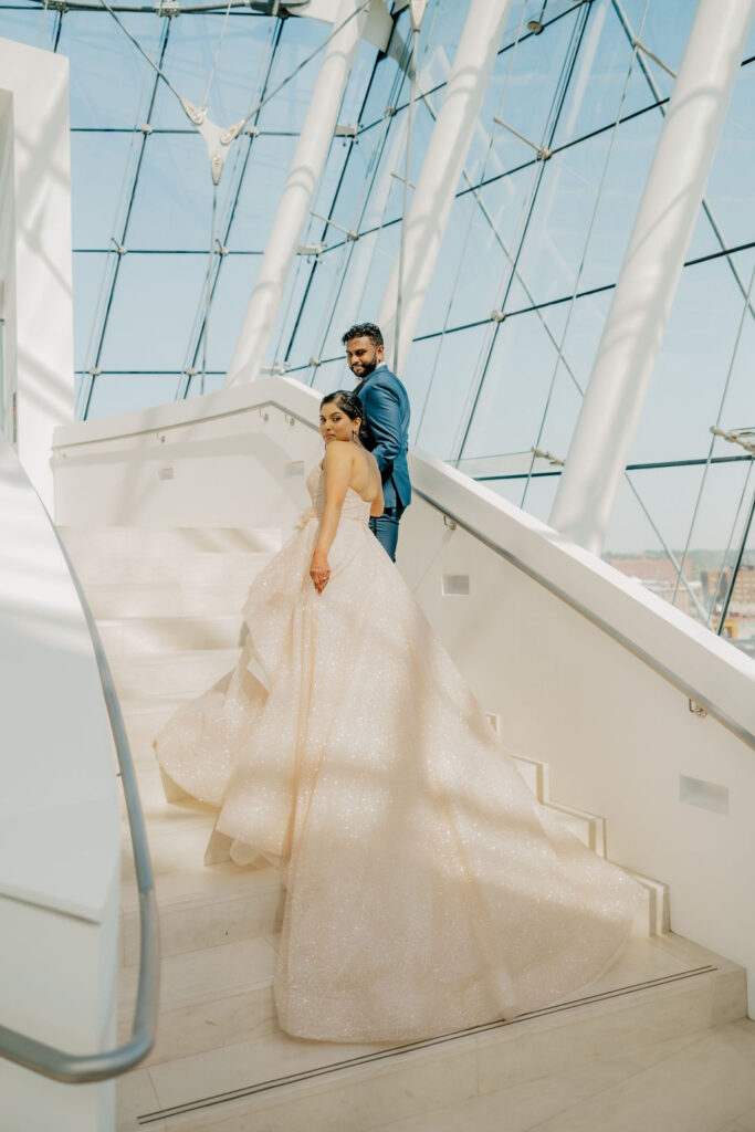 A couple stands on a modern staircase with large windows at the Kauffman Center. The woman is in a flowing, light-colored gown, and the man is in a dark suit. They look back towards the camera. Rent this stunning venue for an unforgettable experience.