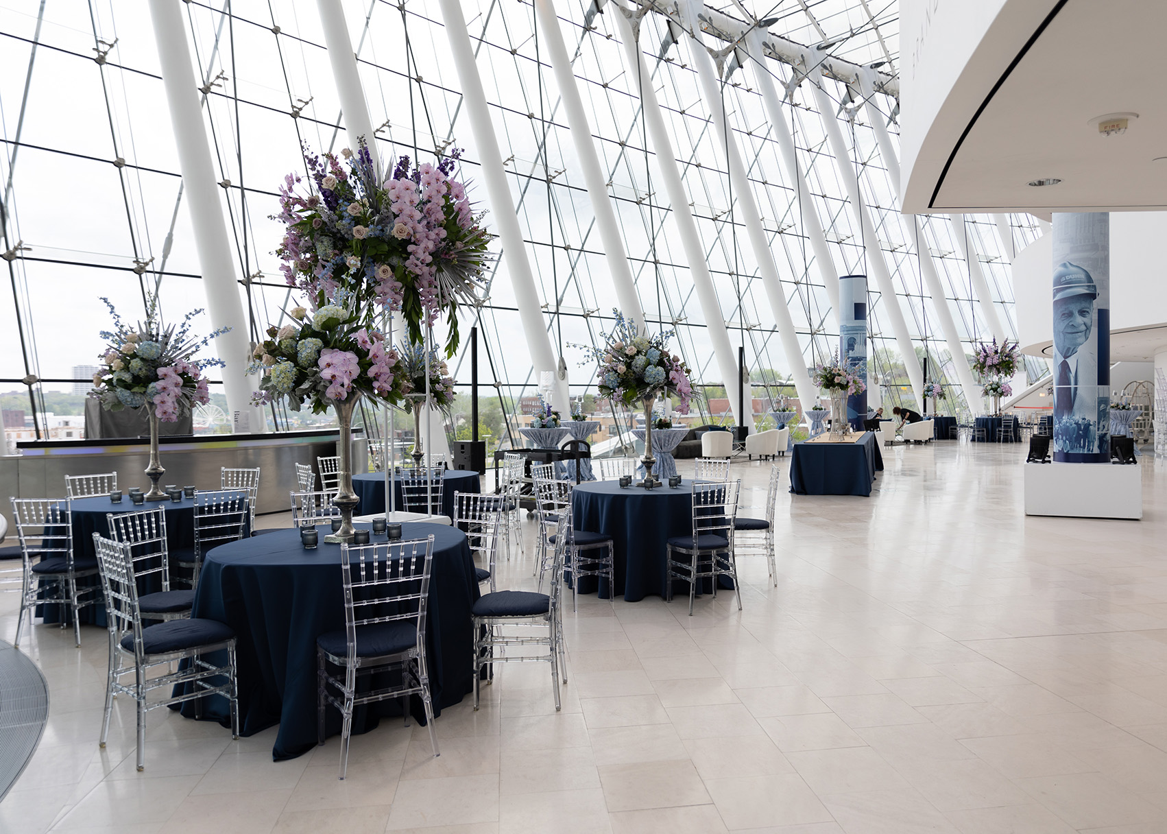 A spacious event hall with high ceilings, large windows, and round tables draped in blue tablecloths. The tables are decorated with tall floral arrangements and surrounded by transparent chairs—perfect for your next gathering when you rent the Kauffman Center.