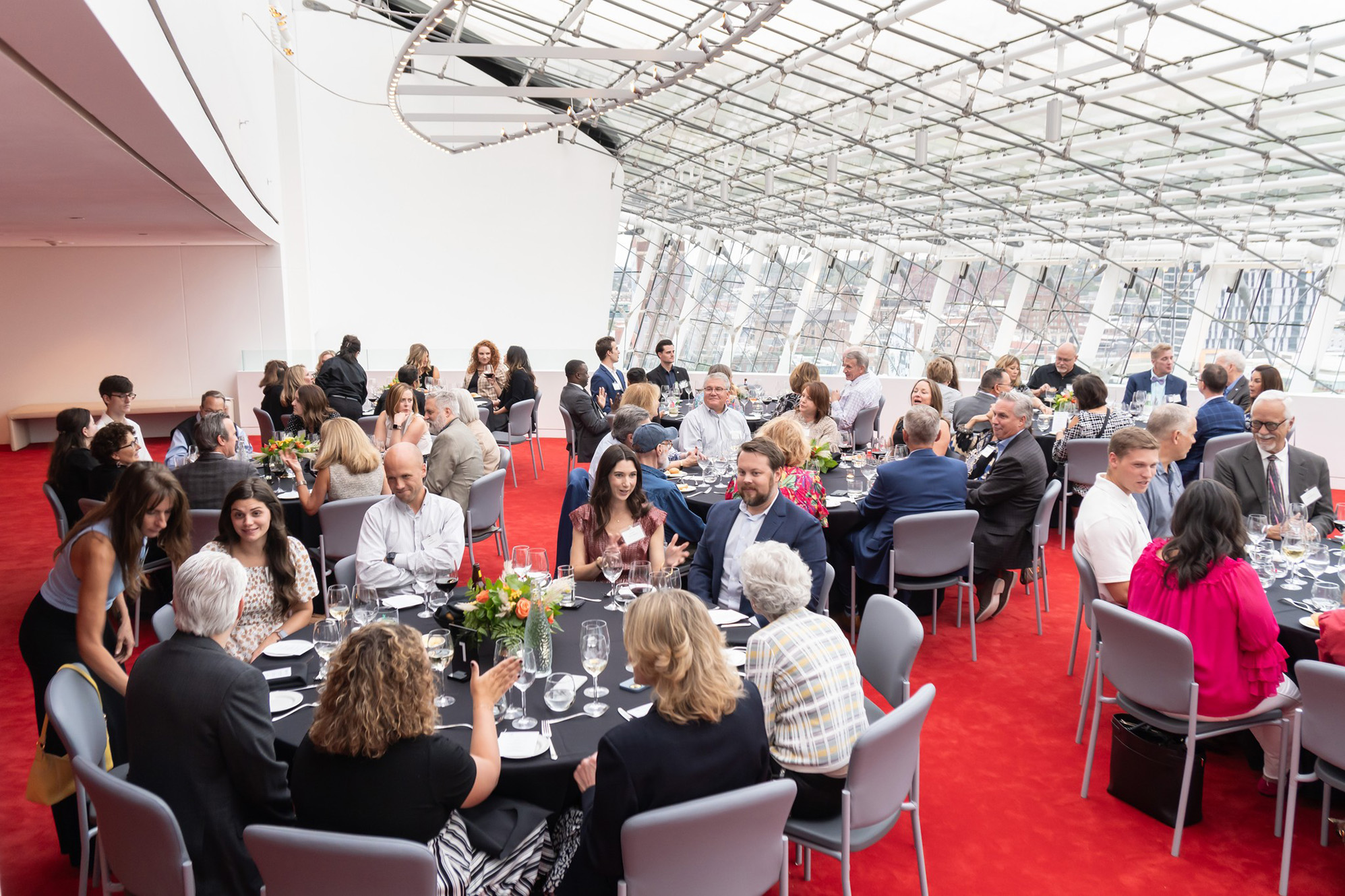 A large group of people in formal attire are seated at round tables for an indoor event in a spacious room with a glass ceiling and red carpet, gathering to discuss business memberships.