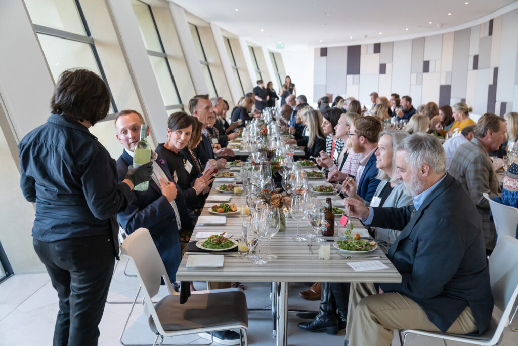 A large group of people, many of whom are likely enjoying the perks of their business memberships, sit at long tables in a modern restaurant. They engage in lively conversation while a staff member pours wine for a guest.