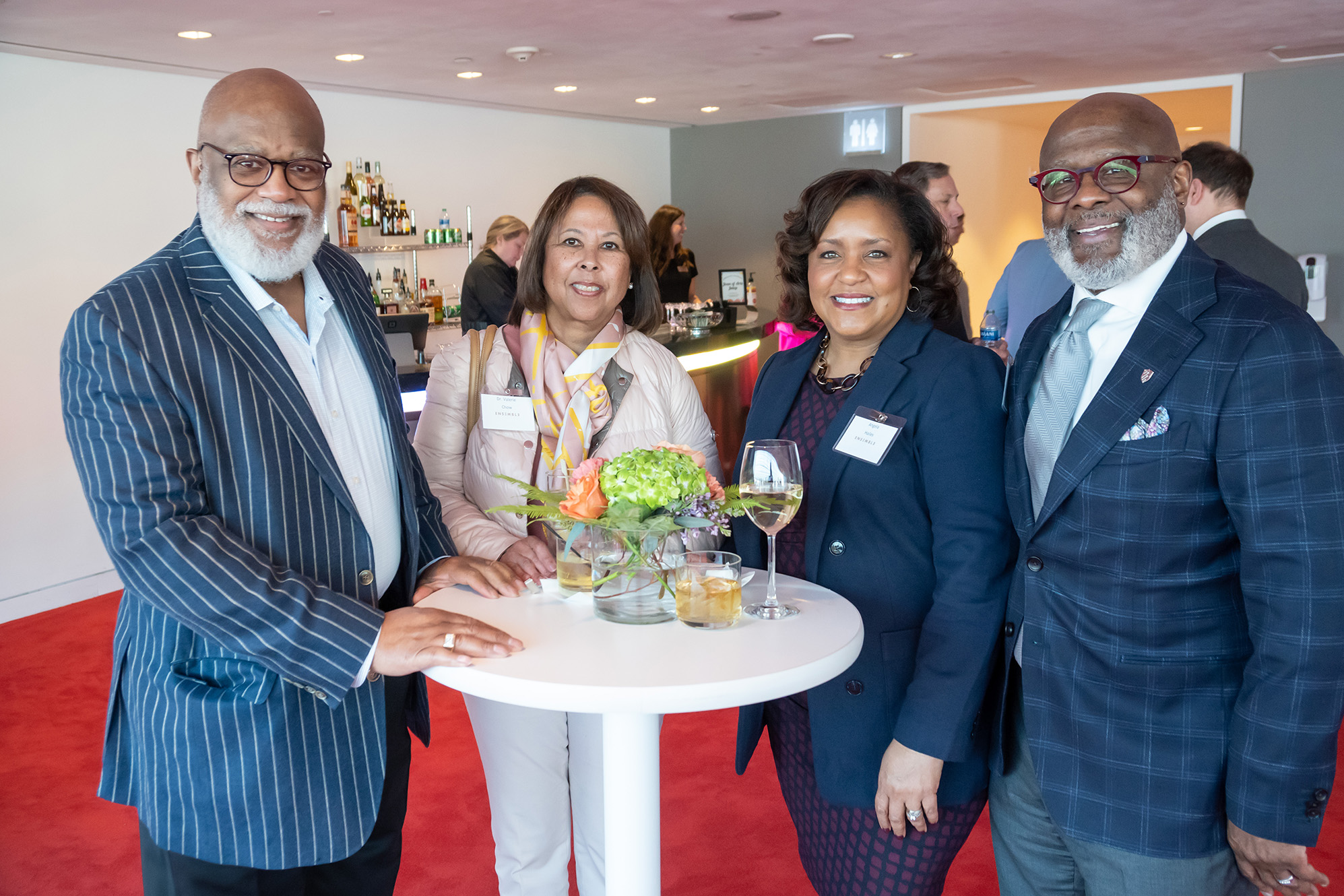 Four people stand around a small table with drinks and flowers. They are dressed in business attire and smiling, showcasing the camaraderie that comes with exclusive business memberships, while a bar and additional people can be seen in the background.