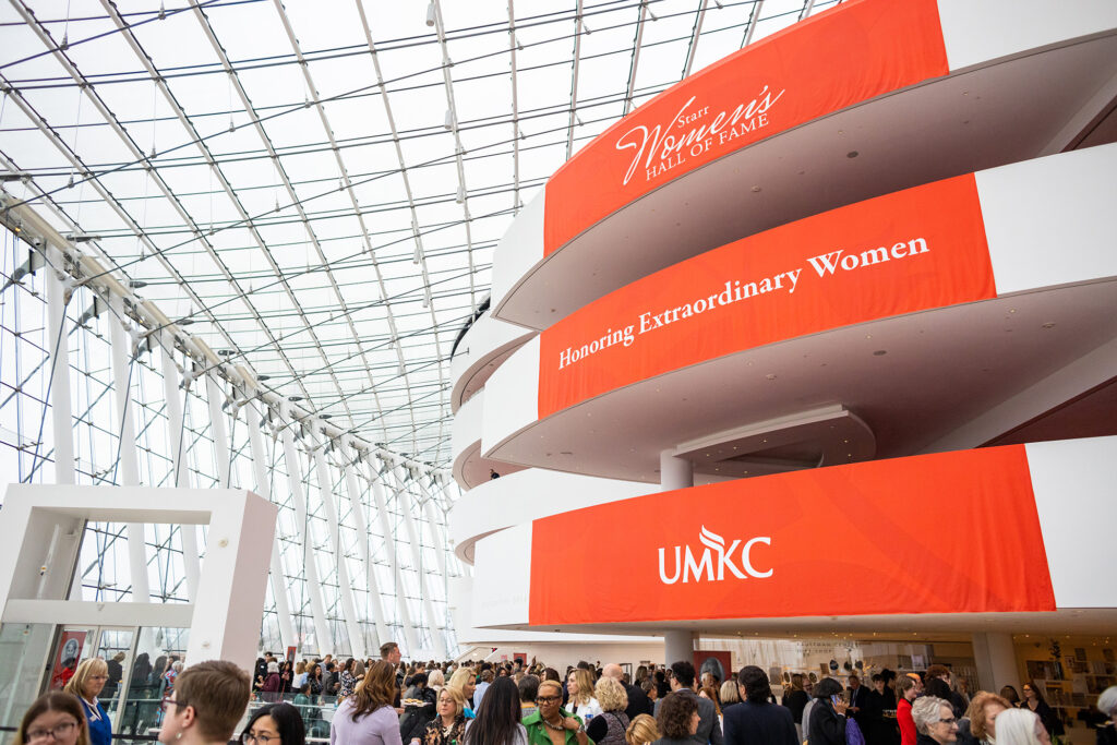 A spacious hall with large red banners that read "Kansas Women's Hall of Fame" and "UMKC" honoring extraordinary women, filled with a crowd of people. For grand events like this, you can rent Kauffman Center to make your celebration unforgettable.