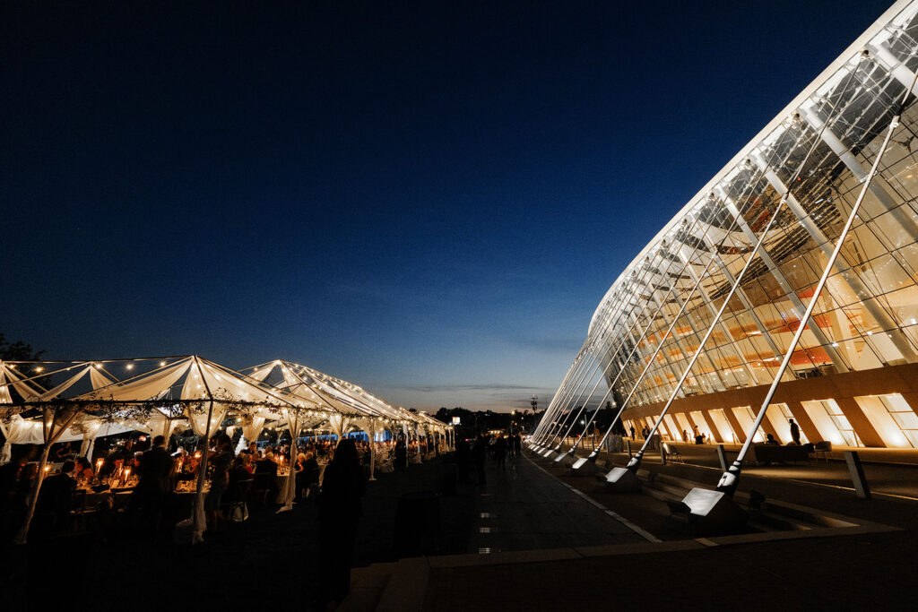 An outdoor event at night under illuminated tents, adjacent to the large, modern Kauffman Center with its stunning glass and steel architecture.