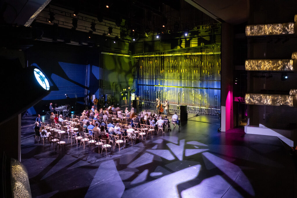 An indoor performance with an audience seated facing a stage area illuminated by colorful lights and geometric patterns on the walls. Musicians are playing on the stage, providing a vibrant backdrop for networking opportunities that come with business memberships.