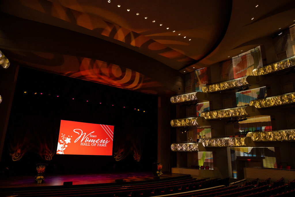 A theater hall with a lit stage displaying the text "Iowa Women's Hall of Fame." The seats and balcony are illuminated, and colorful lights reflect on the walls and ceiling, reminiscent of those you’d find if you rent Kauffman Center for an elegant event.