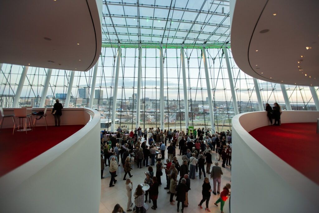 A large group of people gathered in a modern, glass-walled atrium with views of the city skyline in the background, hosted by a prominent Performing Arts Nonprofit in Kansas City.