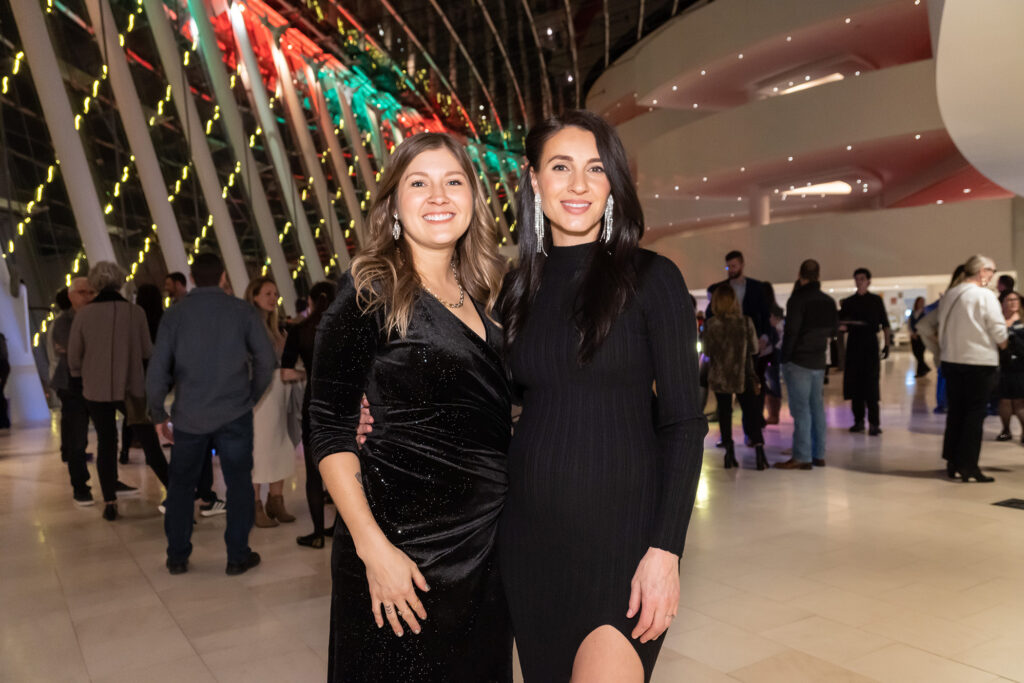 Two women standing indoors, dressed in black outfits—one in a long sleeve velvet dress and the other in a long sleeve dress with a thigh-high slit. People are milling around in the background at an event for a Performing Arts Nonprofit in Kansas City.