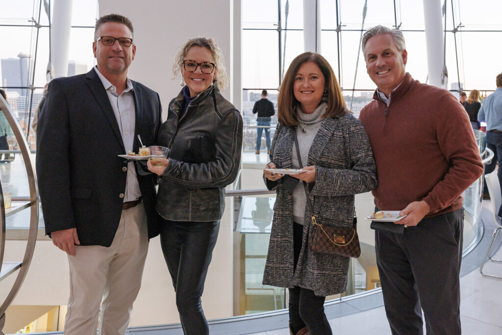 Four people are standing indoors, smiling at the camera, holding plates of food. Casually dressed, they are in a modern setting with large windows in the background—a snapshot from a Performing Arts Nonprofit event in Kansas City.