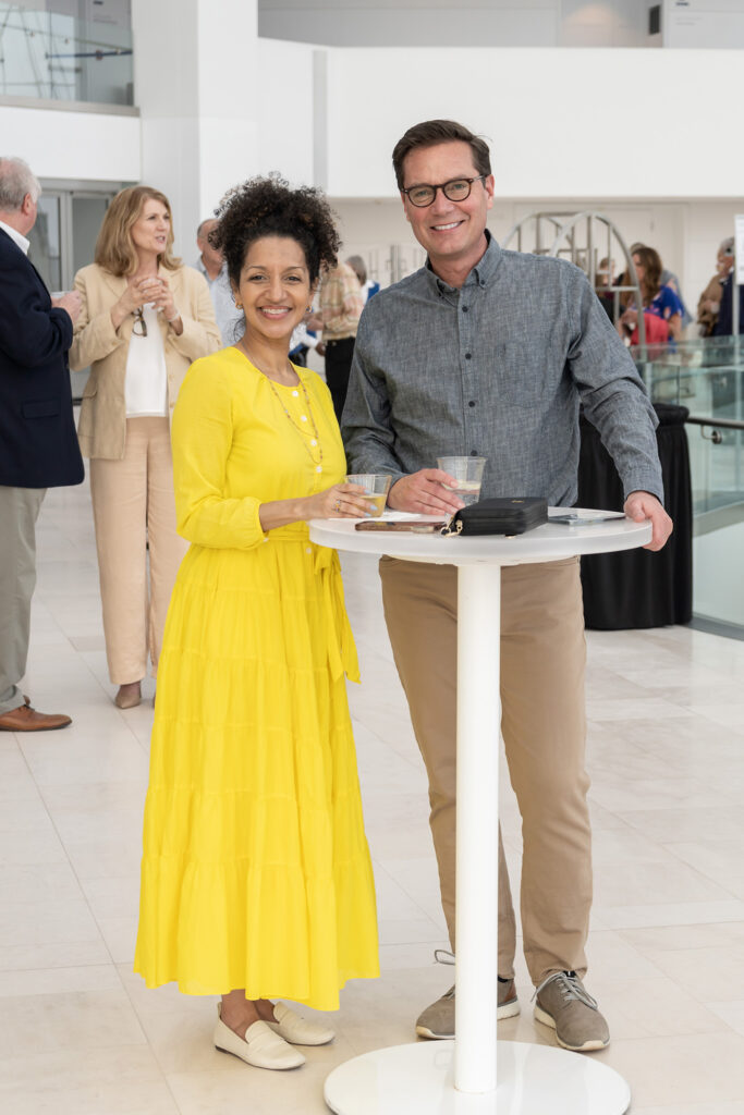 A woman in a yellow dress and a man in a gray shirt stand together, smiling, at a high-top table, holding drinks. Others are seen in conversation in the background. It appears to be an event for a Performing Arts Nonprofit in Kansas City.