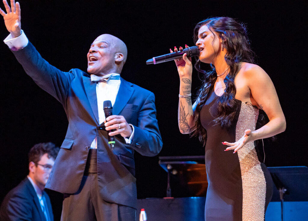 Two singers perform on stage, holding microphones. One is in a tuxedo, raising his hand, and the other is in a black dress, singing beautifully. Backed by a skilled pianist, this enchanting scene showcases the talent supported by our Performing Arts Nonprofit in Kansas City.