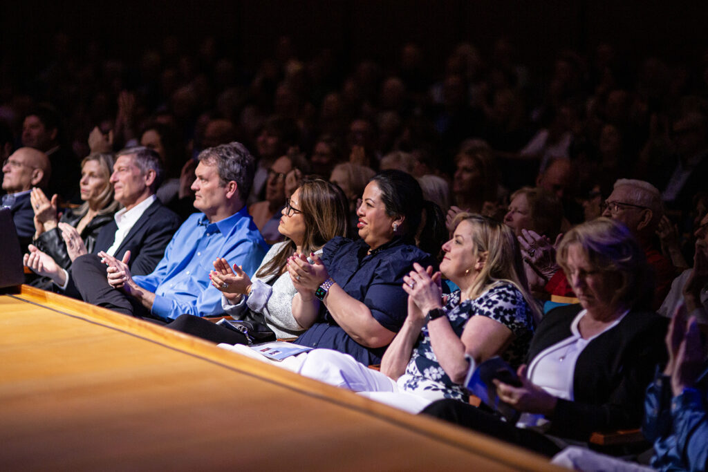 A group of people sitting in an auditorium, attending a presentation by a Performing Arts Nonprofit in Kansas City, applauding with expressions of appreciation on their faces.