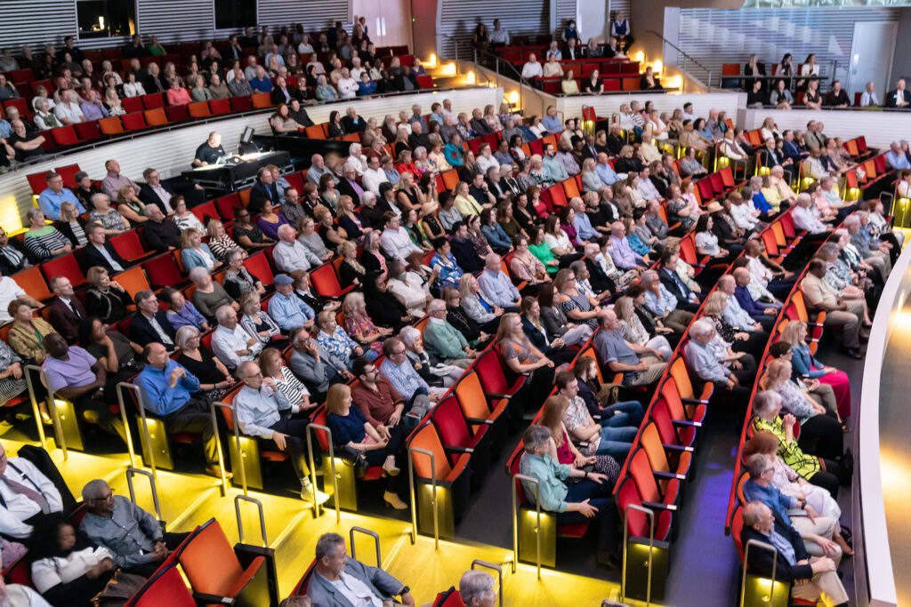 A large audience seated in a modern auditorium, watching an event or performance hosted by a Performing Arts Nonprofit in Kansas City, with people filling most of the red and orange chairs.