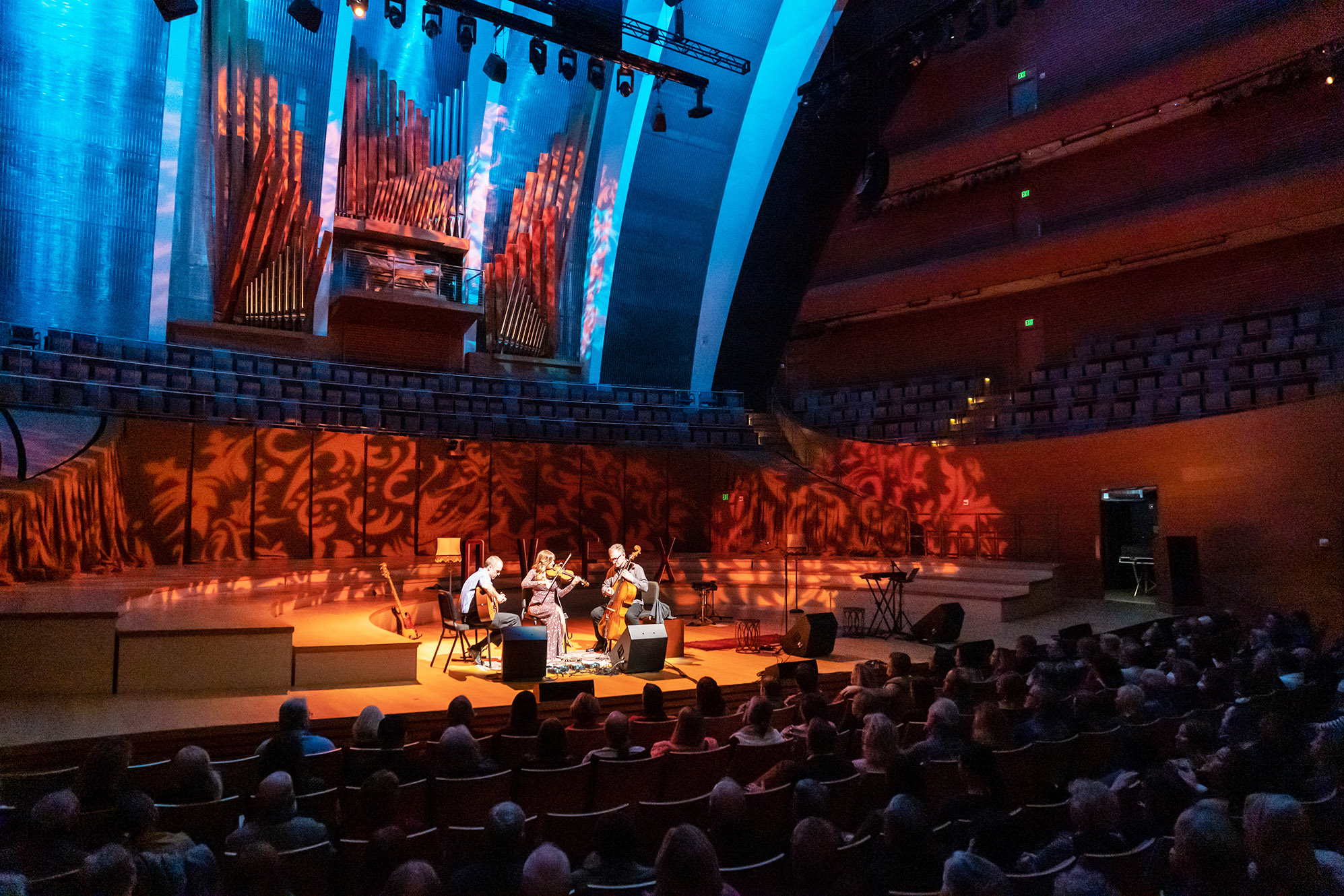 A string quartet from a renowned performing arts nonprofit in Kansas City performs on stage in front of an audience in a concert hall with vivid light projections and an organ in the background.